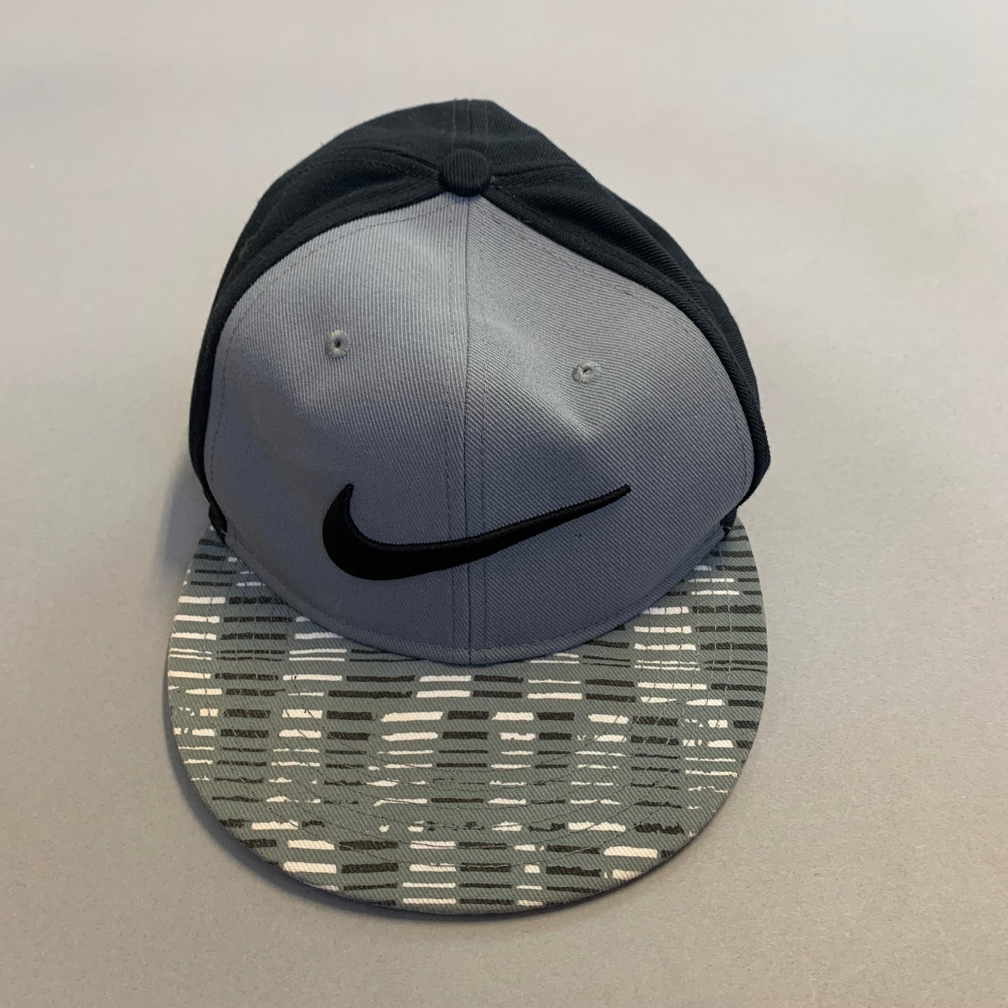 Grey cap with a black logo and patterned brim on a flat surface.