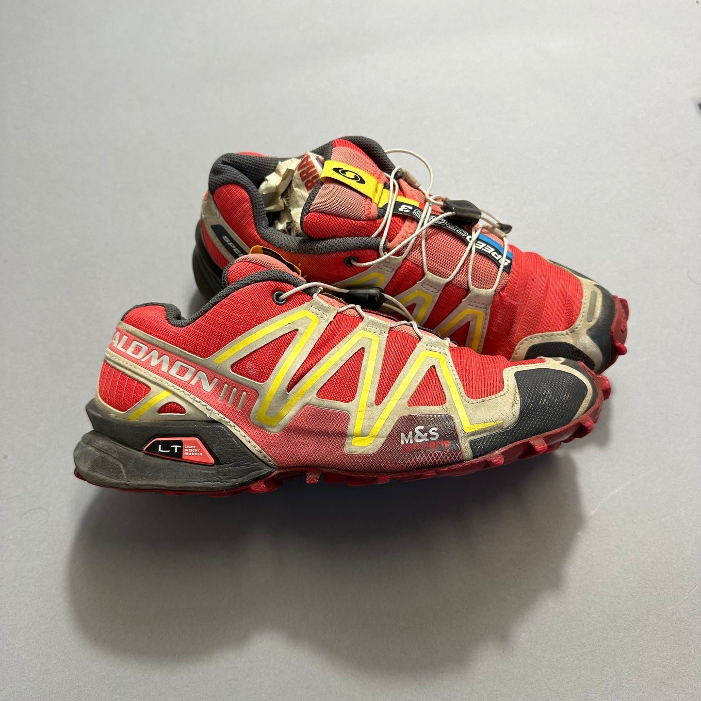 A pair of red and yellow trail running shoes on a gray background.