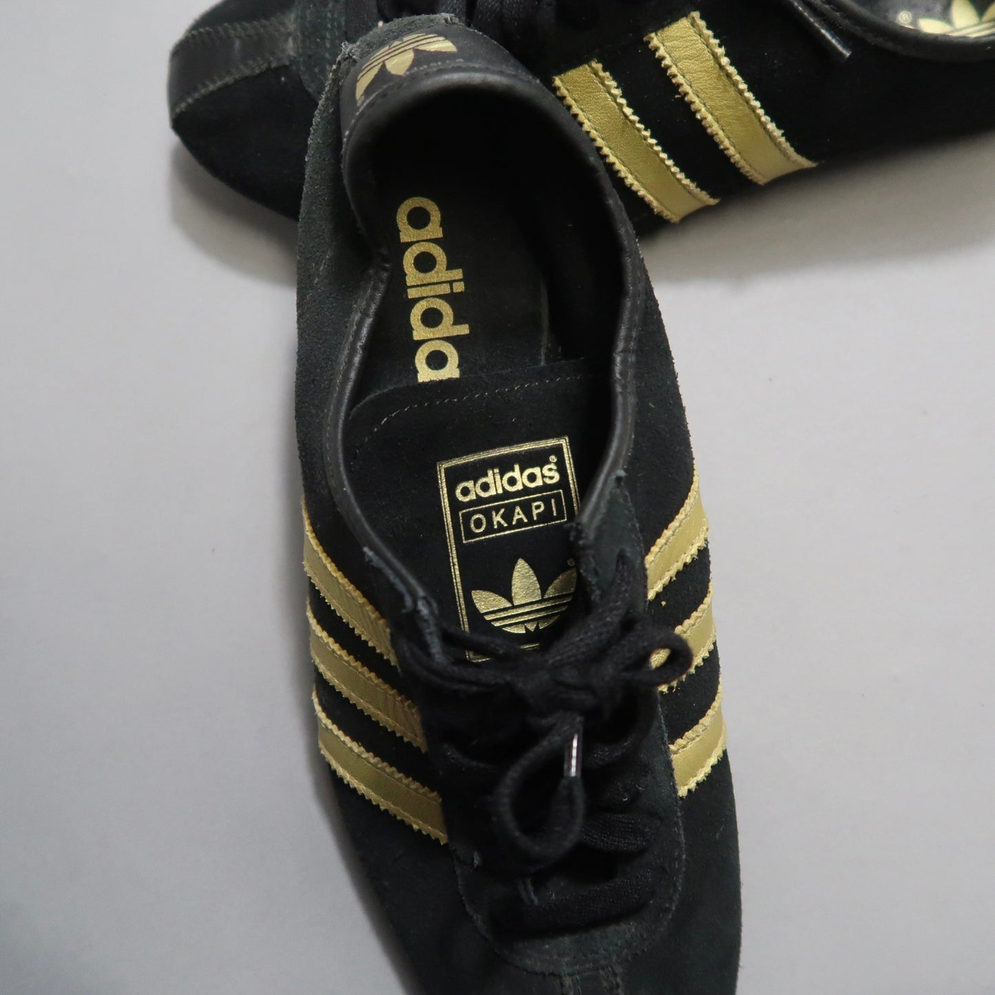 Black Adidas Okapi sneakers with gold stripes on a gray background.