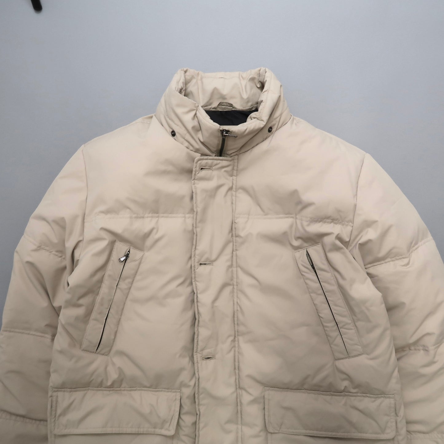 Beige puffer jacket with high collar and zippered pockets.