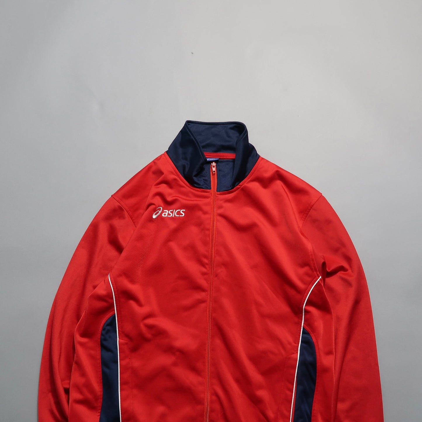 Red Asics sports jacket with navy accents on a gray background.