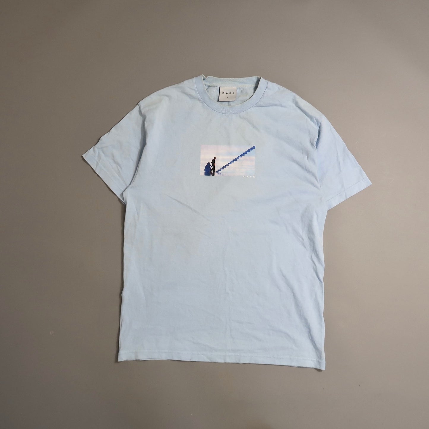Light blue T-shirt with a small print of a person climbing stairs.