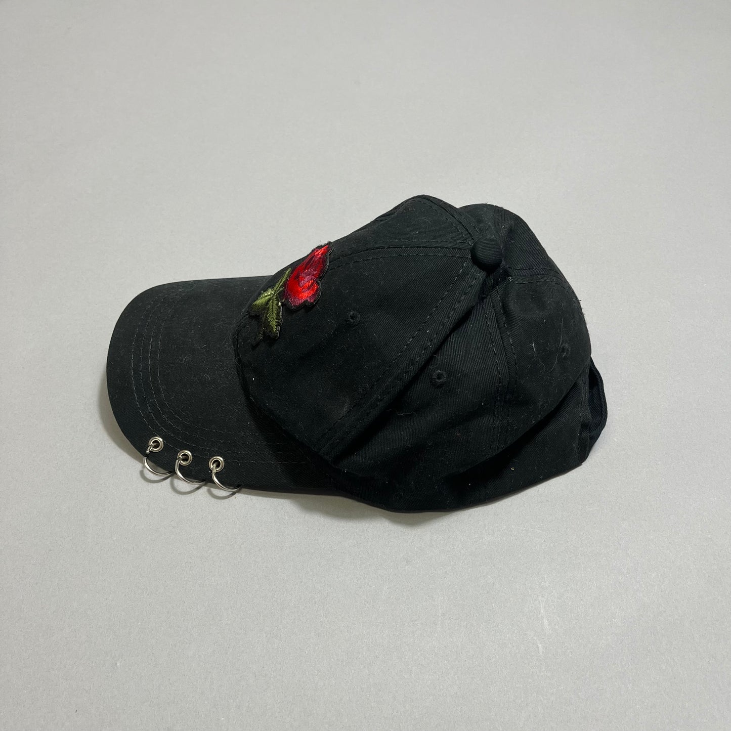 Black cap with a red rose patch and three metal rings on the brim.
