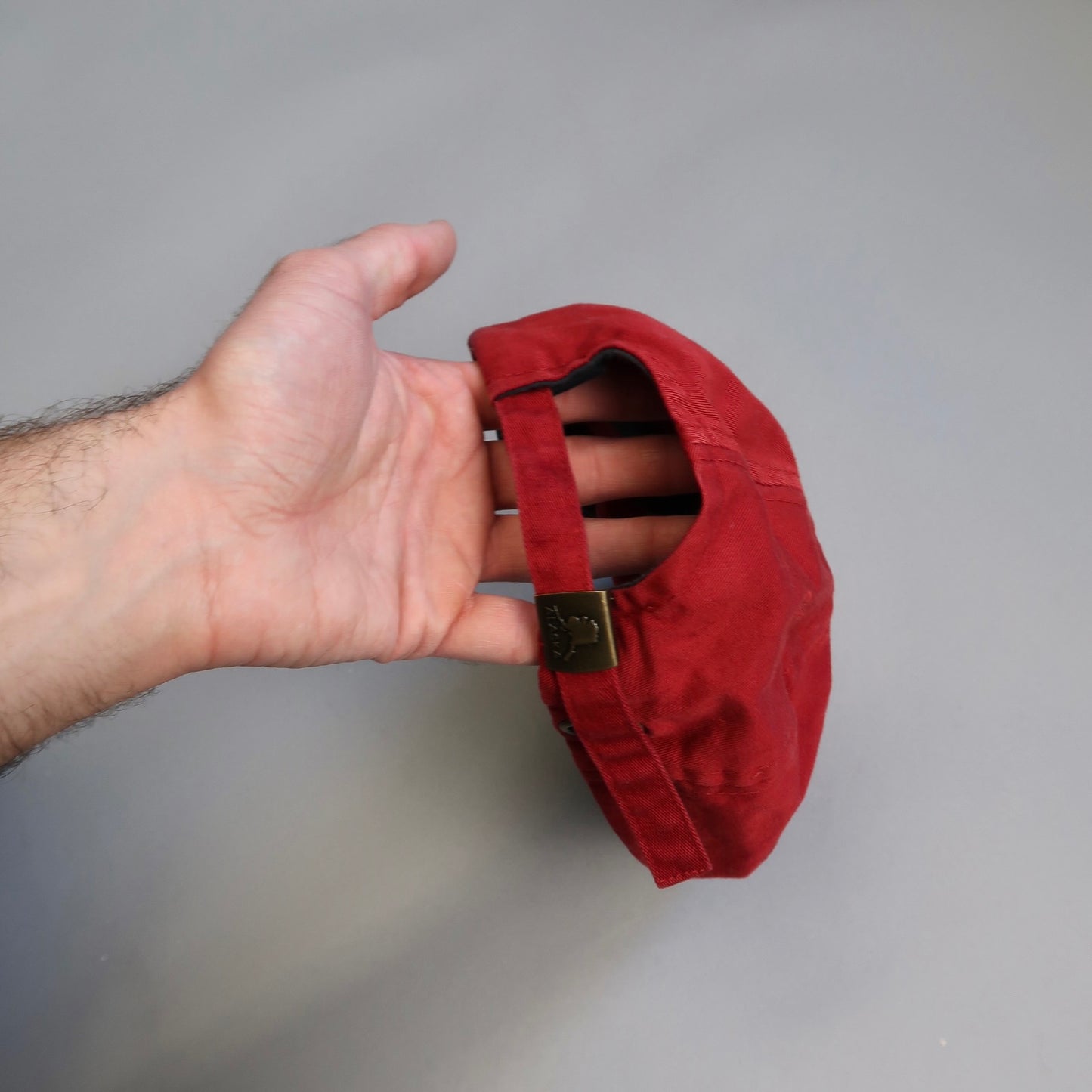 Hand holding a small red cap with an adjustable strap.