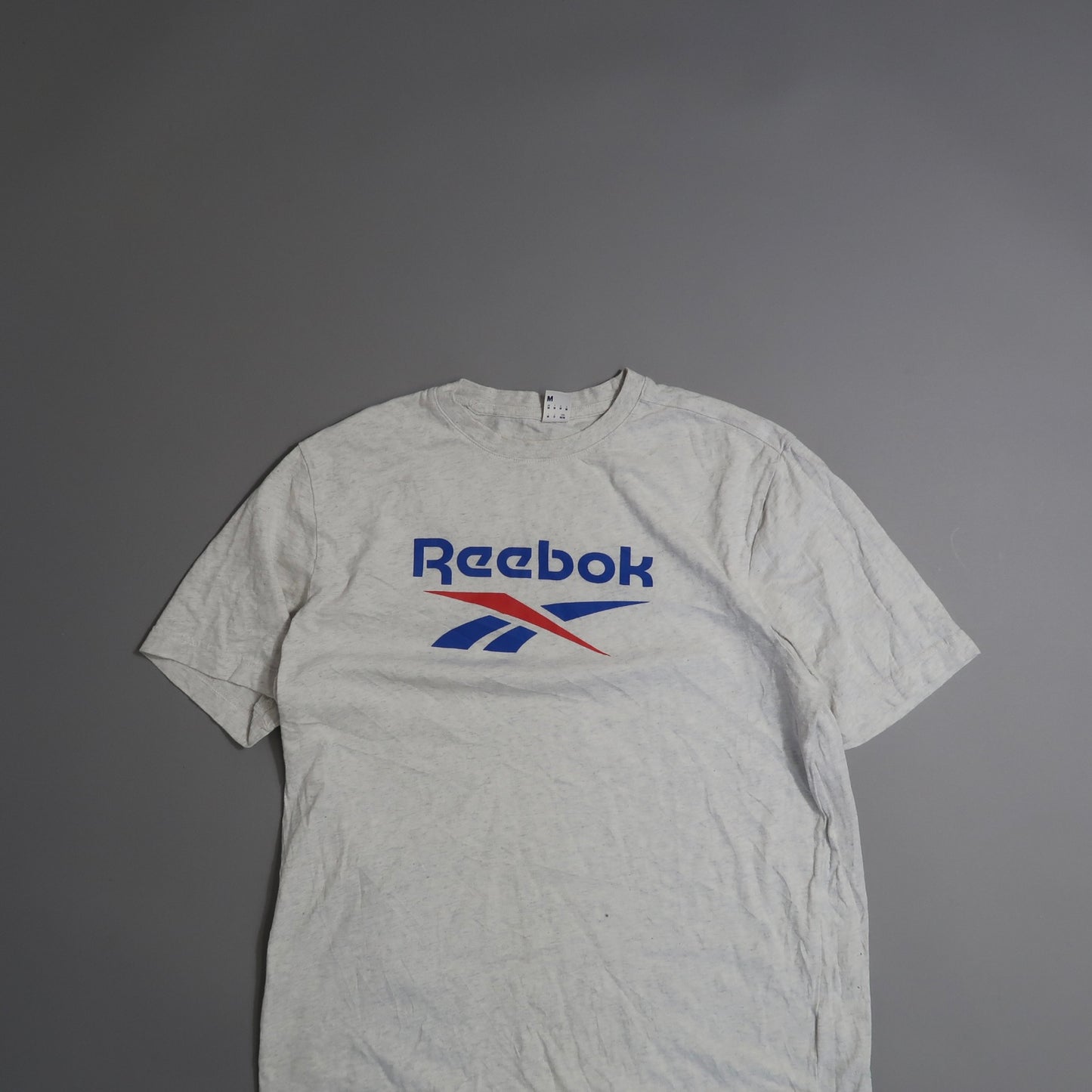 Gray Reebok t-shirt with blue and red logo on a gray background.