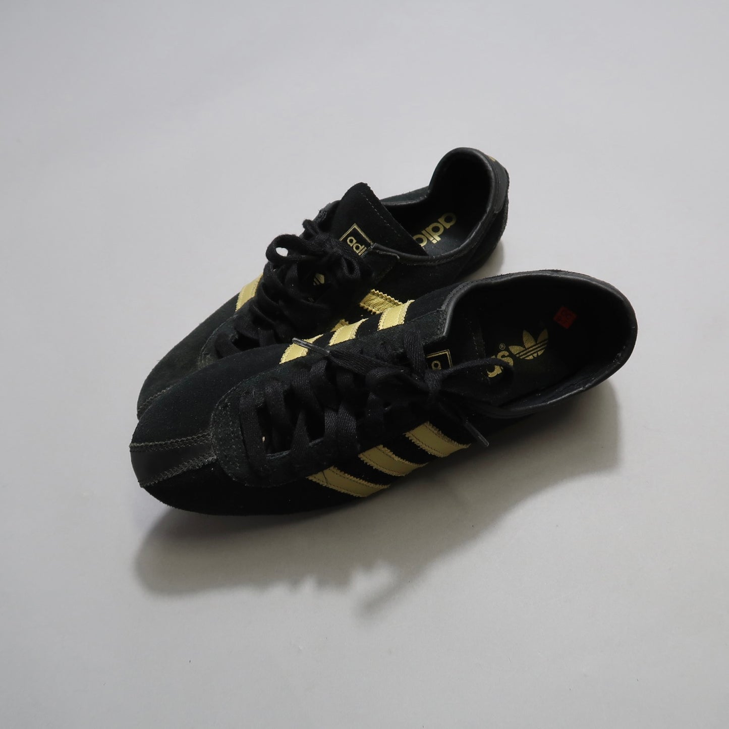 Black sneakers with gold stripes on a light gray background.