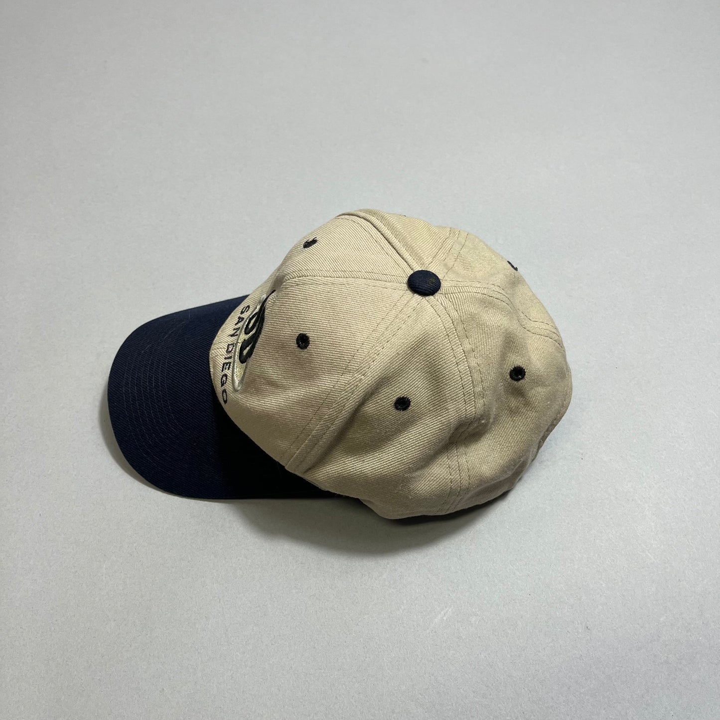 Tan baseball cap with a navy blue brim on a neutral background.