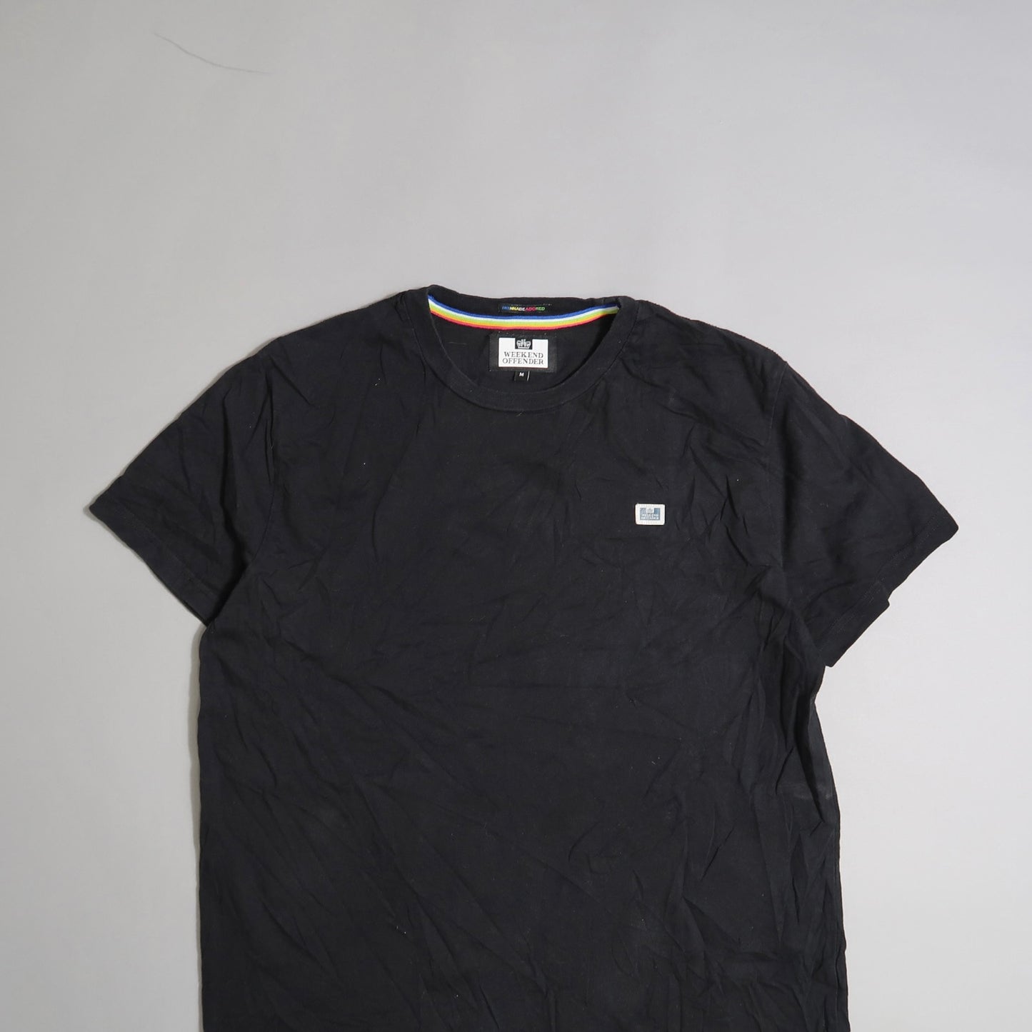 Black T-shirt with a small chest logo on a light gray background.