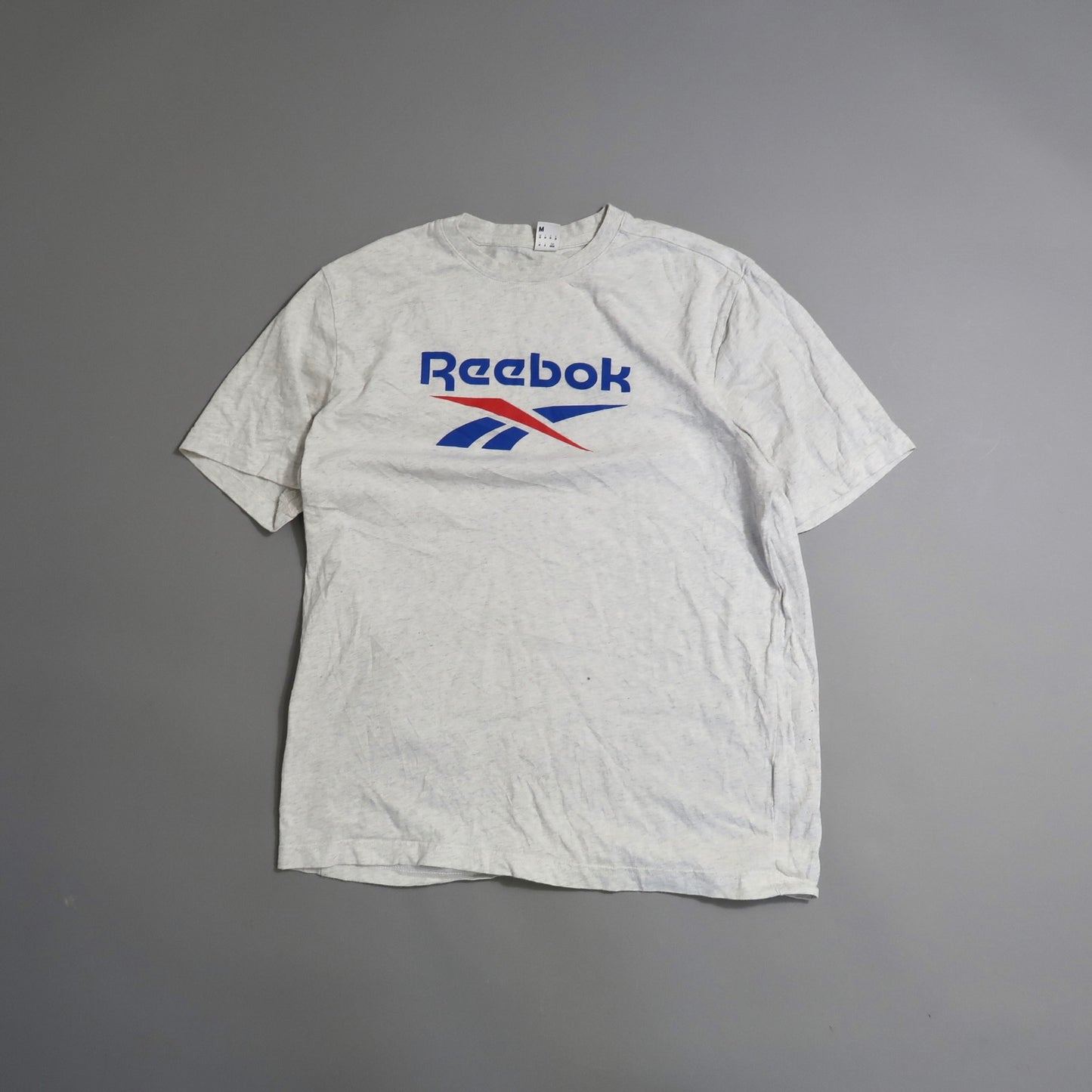 White Reebok T-shirt with blue and red logo on gray background.