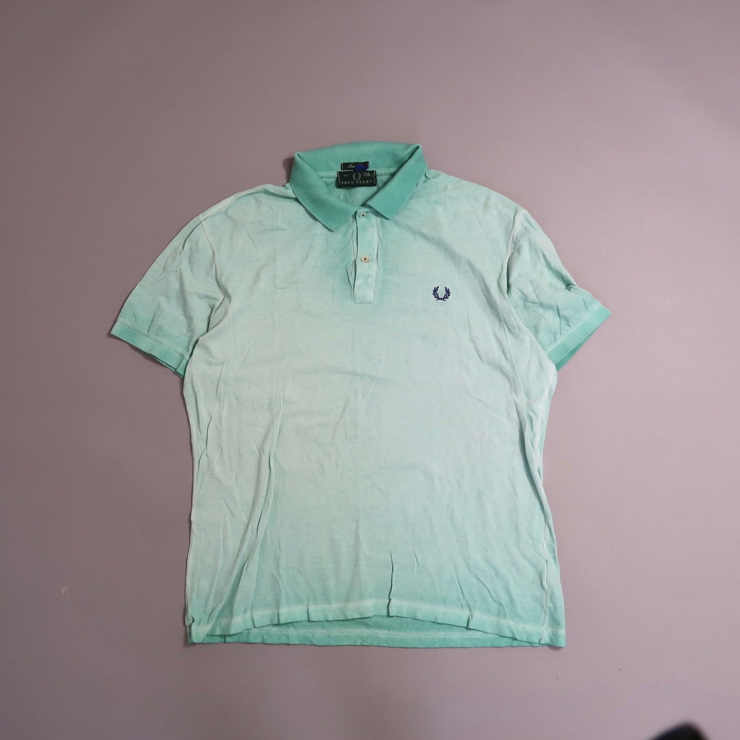 Light green polo shirt with dark logo on chest, laid flat on gray surface.