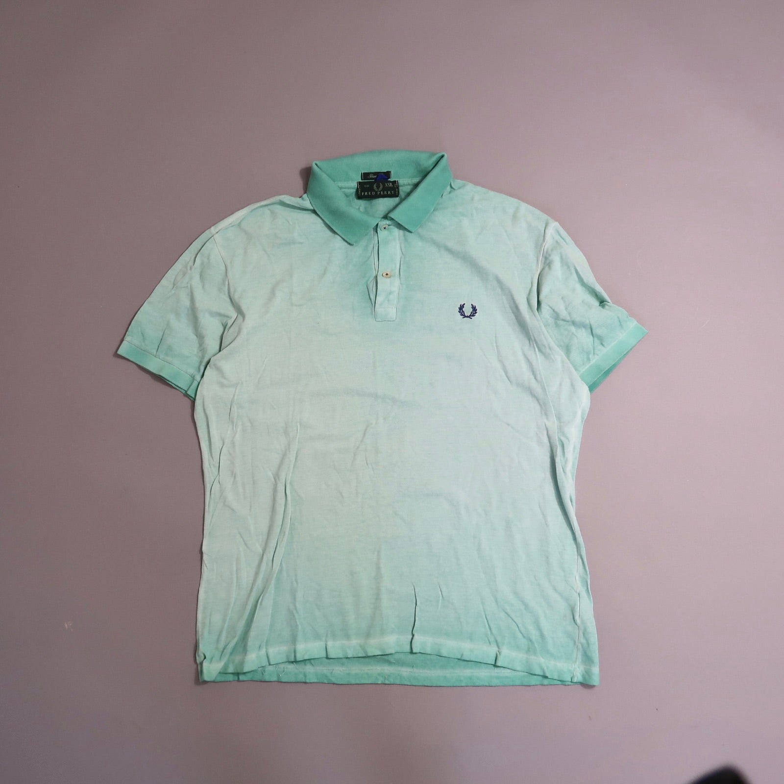 Light green polo shirt with dark logo on chest, laid flat on gray surface.