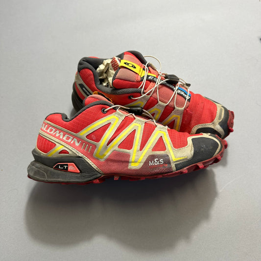 A pair of red and yellow trail running shoes on a gray background.