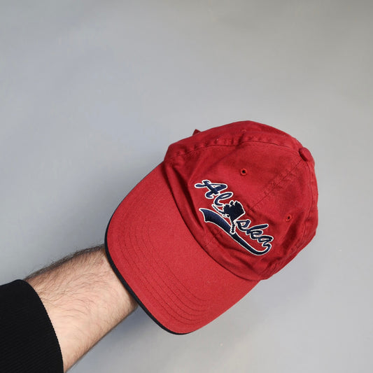 Red cap with nullAlaskanull embroidery, held by a person's hand.