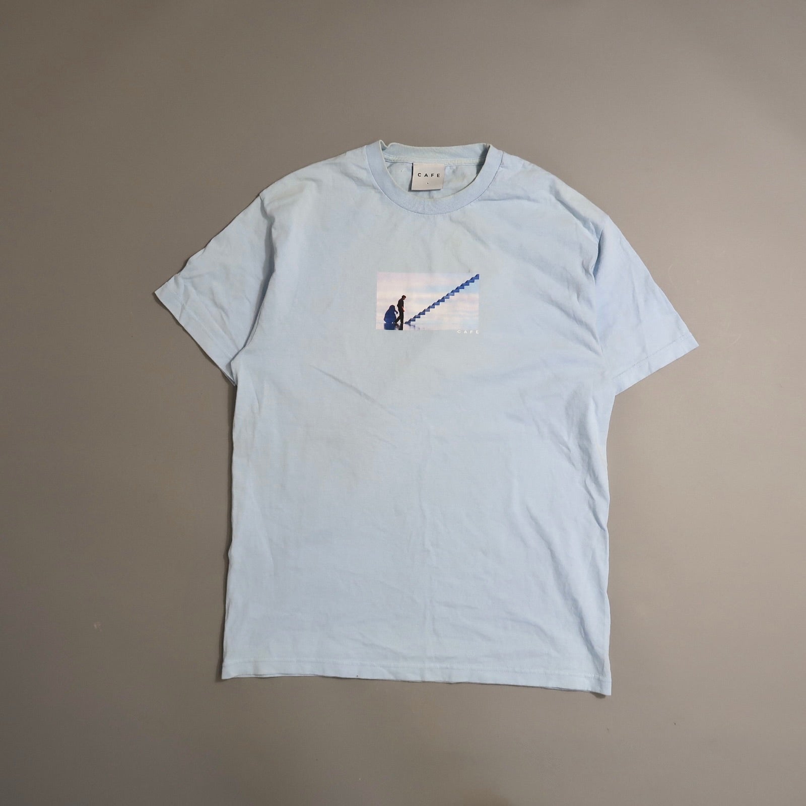 Light blue T-shirt with a small print of a person climbing stairs.