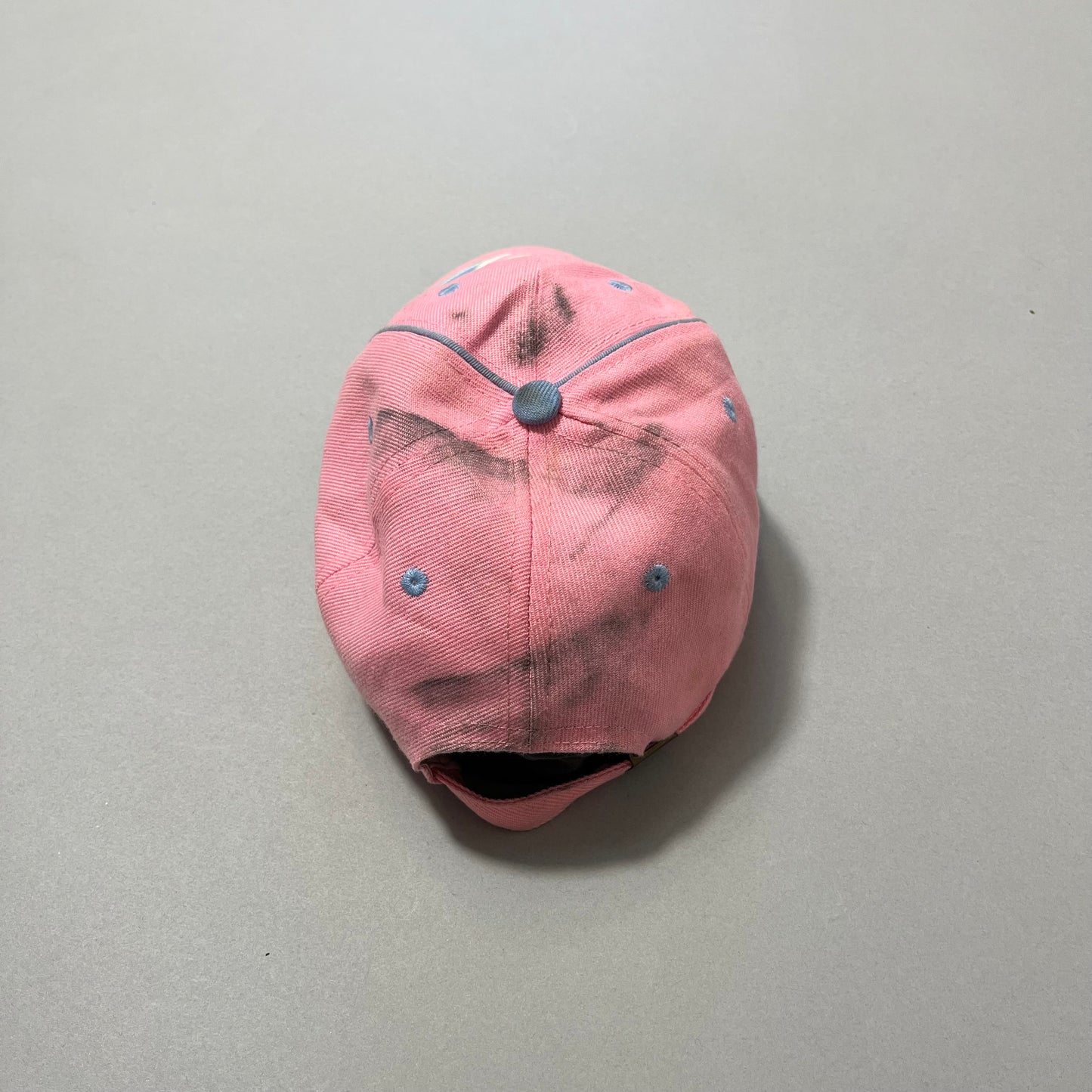 Worn pink cap with blue eyelets and a button, placed on a gray background.