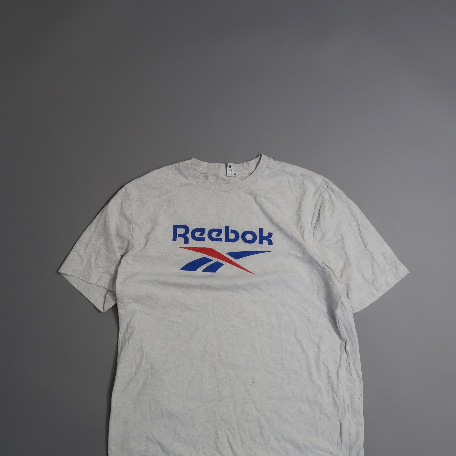 Gray Reebok t-shirt with blue and red logo on a gray background.