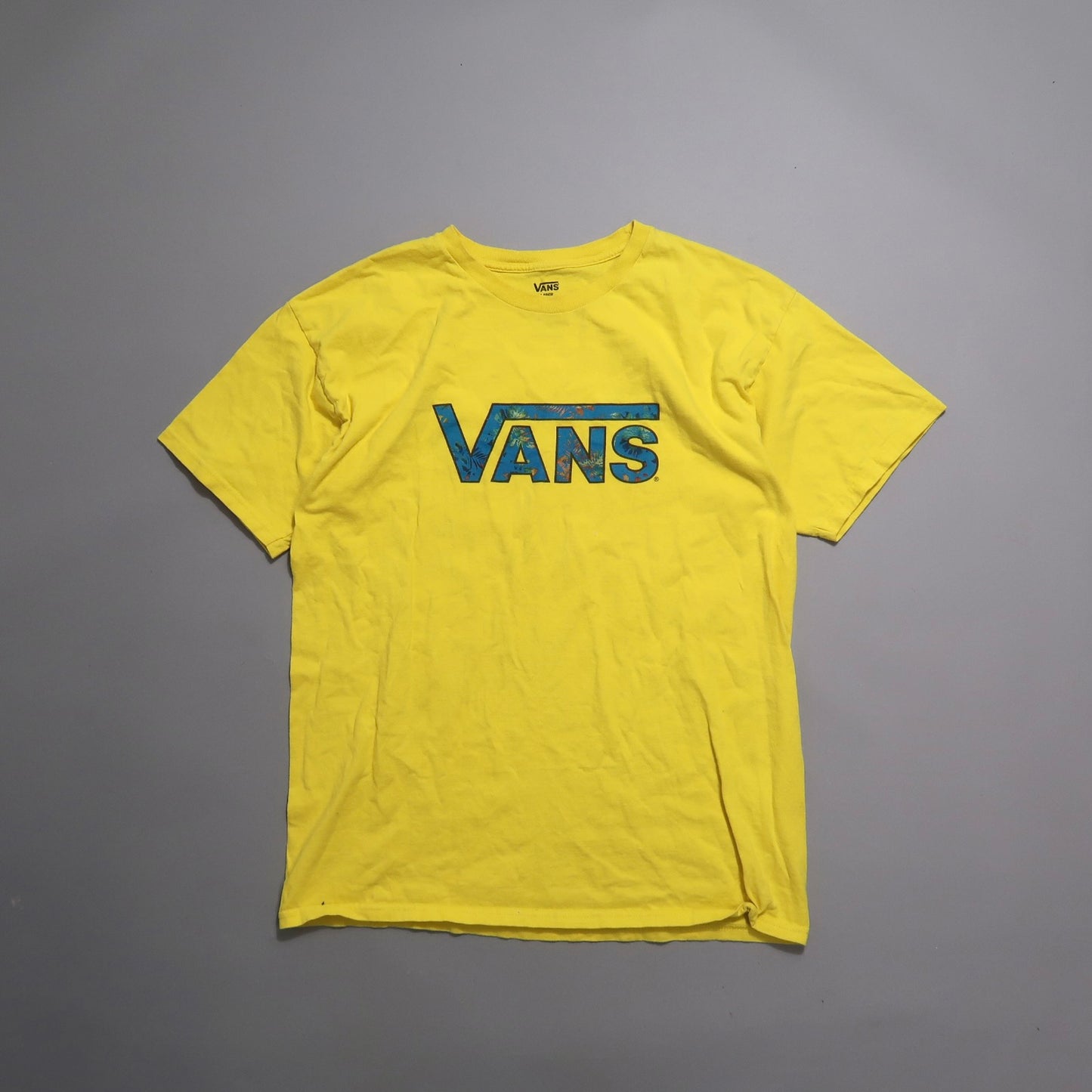 Yellow Vans t-shirt with blue floral logo on a gray background.