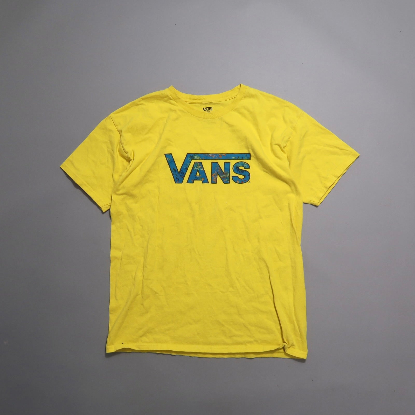 Yellow Vans t-shirt with blue floral logo on a gray background.