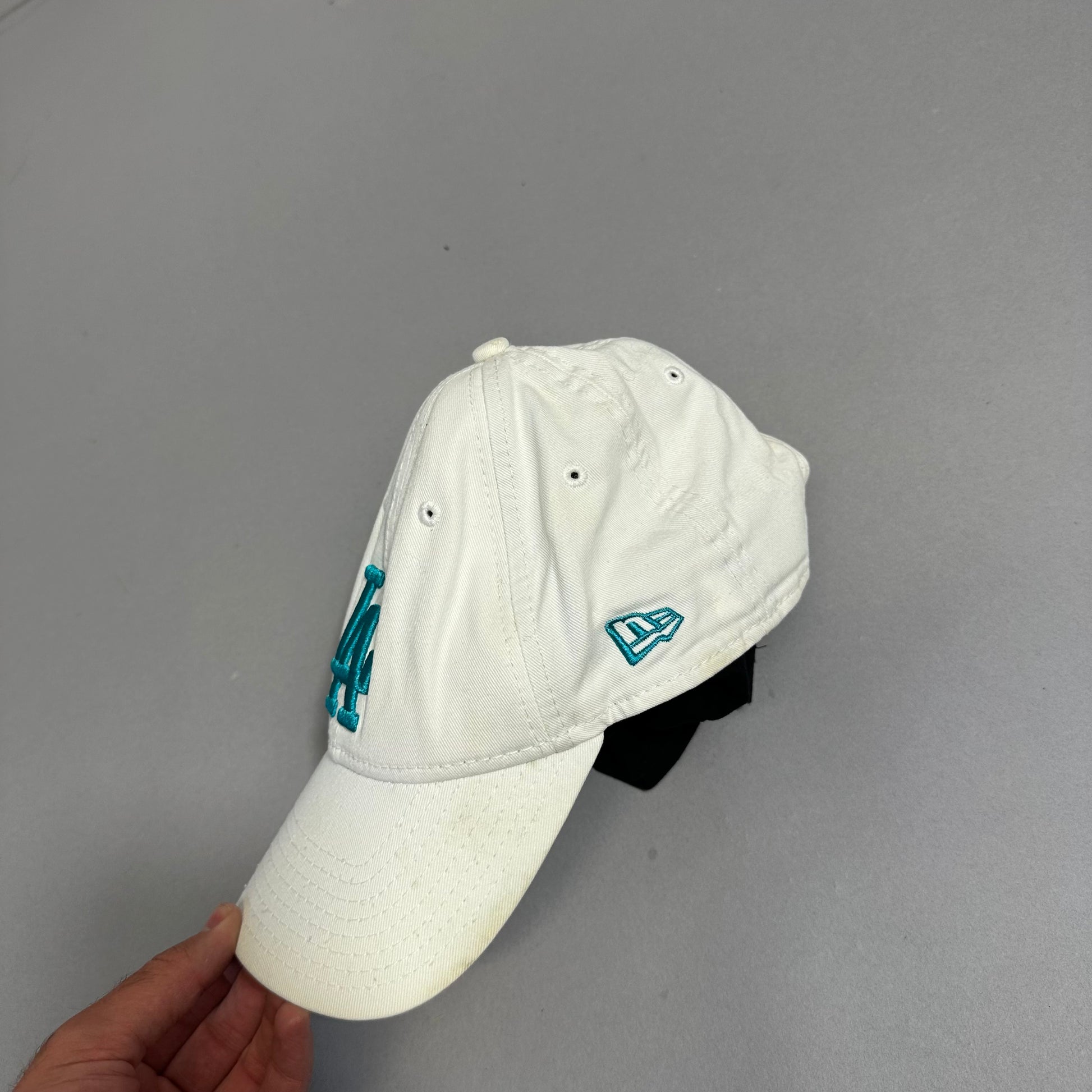 White baseball cap with teal logo, held by a hand.