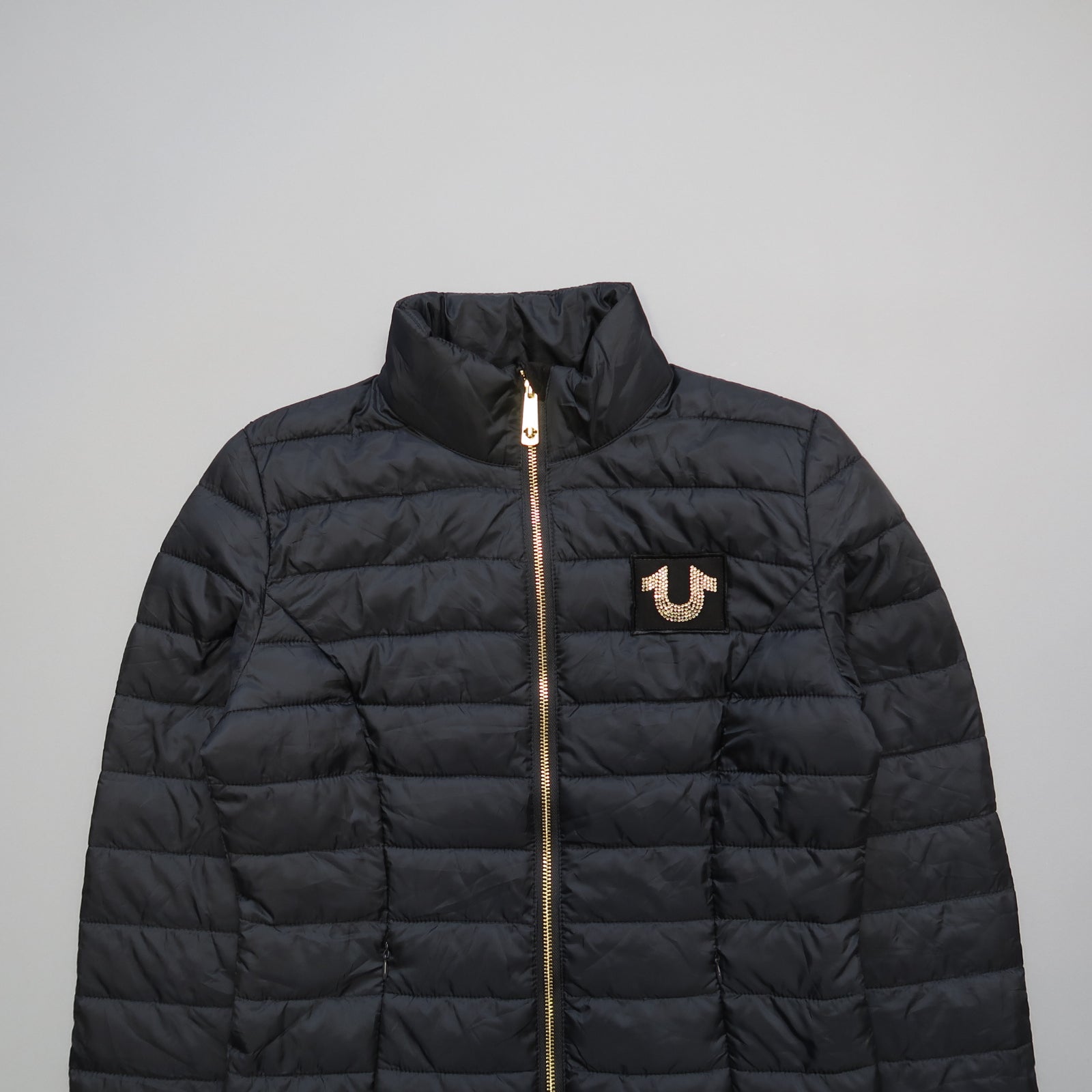 Black quilted jacket with gold zipper and logo on chest.